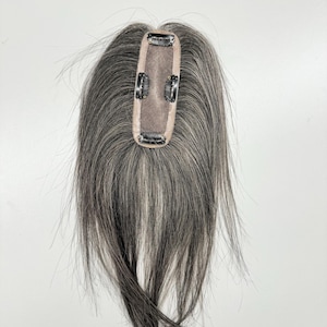 May include: A hair topper with a rectangular base and three clips. The hair is a mix of gray and dark brown, with a natural-looking texture. The base is a light beige color.