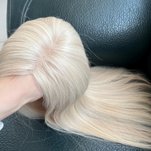 May include: A straight, platinum blonde wig featuring a visible scalp part with a skin-toned base. The hair is long, smooth, and light-colored, showcasing a natural-looking parting area that transitions into sleek, flowing strands of synthetic hair.