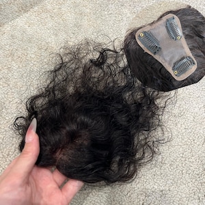 May include: A dark brown hair topper with a wavy texture and a natural-looking scalp base. The underside features a beige mesh foundation with three metal pressure-sensitive clips attached for secure placement on the head.