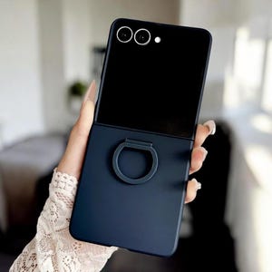 Puede incluir: Una funda de teléfono azul marino con un soporte para anillos. La funda está diseñada para un teléfono plegable, con una sección superior negra y una sección inferior azul marino. El soporte para anillos es de color azul marino a juego.