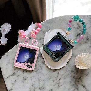 Peut inclure: Deux smartphones pliables, un rose et un vert, sont présentés. Le téléphone rose a un bord blanc et un charm floral. Le téléphone vert a une dragonne perlée. Les deux téléphones affichent l'heure 21:24 sur leurs écrans.