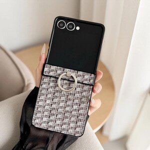 Puede incluir: Funda de teléfono negra con un patrón tejido gris y blanco y un soporte de anillo plateado. El teléfono está abierto, revelando las lentes de la cámara.