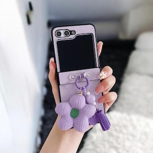 May include: A lilac phone case with a corduroy texture, featuring a black screen and camera lenses. A flower-shaped charm with a green centre and a purple tassel hangs from the case. The case is held in a hand.