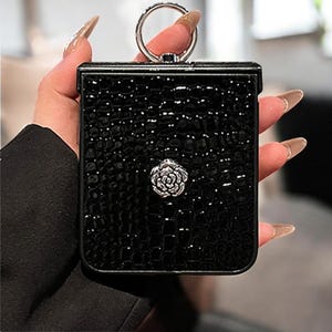 Può includere: Pochette rettangolare nera con un motivo strutturato a effetto coccodrillo. La pochette presenta un manico ad anello color argento e un'applicazione a forma di rosa argentata. La borsa è tenuta da una persona.