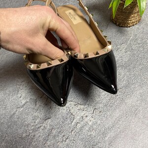 May include: Black patent leather flats with pointed toes and a slingback strap. The shoes feature a beige trim adorned with gold-colored pyramid studs. The Valentino logo is visible on the insole.