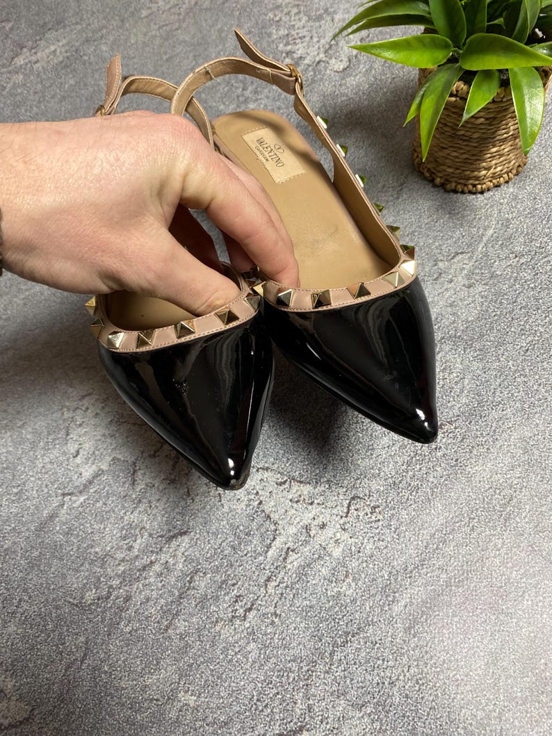 May include: Black patent leather flats with pointed toes and a slingback strap. The shoes feature a beige trim adorned with gold-colored pyramid studs. The Valentino logo is visible on the insole.