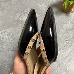 May include: Pair of black patent leather high heels with pointed toes. The shoes have a nude-colored strap with gold-colored pyramid studs. The heels are held in a person's hand.