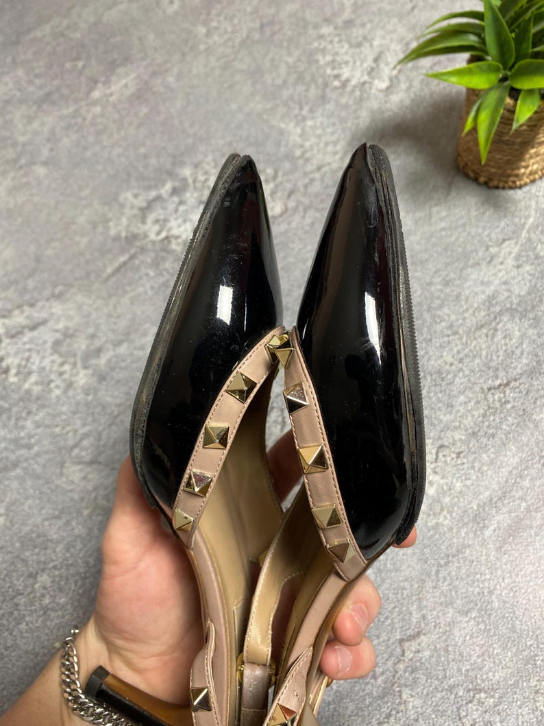 May include: Pair of black patent leather high heels with pointed toes. The shoes have a nude-colored strap with gold-colored pyramid studs. The heels are held in a person's hand.