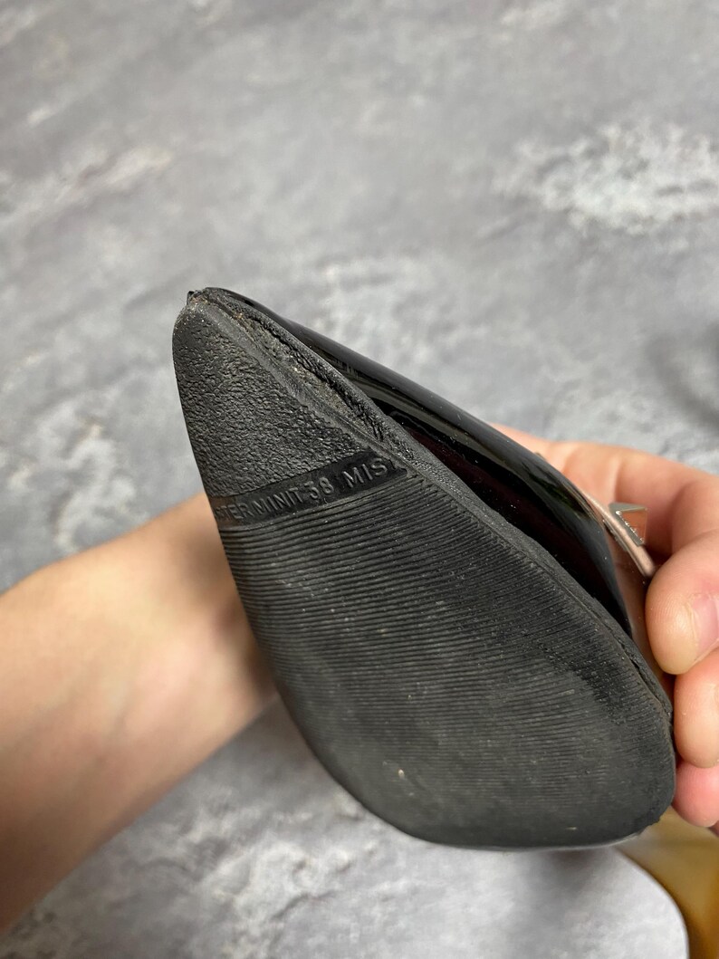 May include: Black patent leather shoe with a textured sole. The shoe's sole has the text "TERMINIT 38 MIS". The shoe is held by a person.