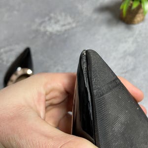 May include: Close-up of a black pointed-toe shoe. The sole is textured with a ridged pattern. The shoe is held by a person's hand. A second shoe is visible in the background.