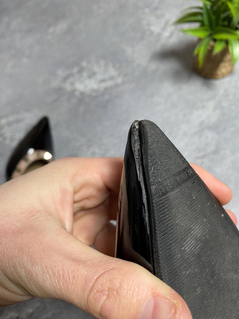 May include: Close-up of a black pointed-toe shoe. The sole is textured with a ridged pattern. The shoe is held by a person's hand. A second shoe is visible in the background.