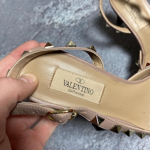 May include: Close-up of a pair of light pink Valentino Garavani sandals. The sandals feature small gold-colored pyramid studs and a thin ankle strap. The Valentino logo is visible on the insole.