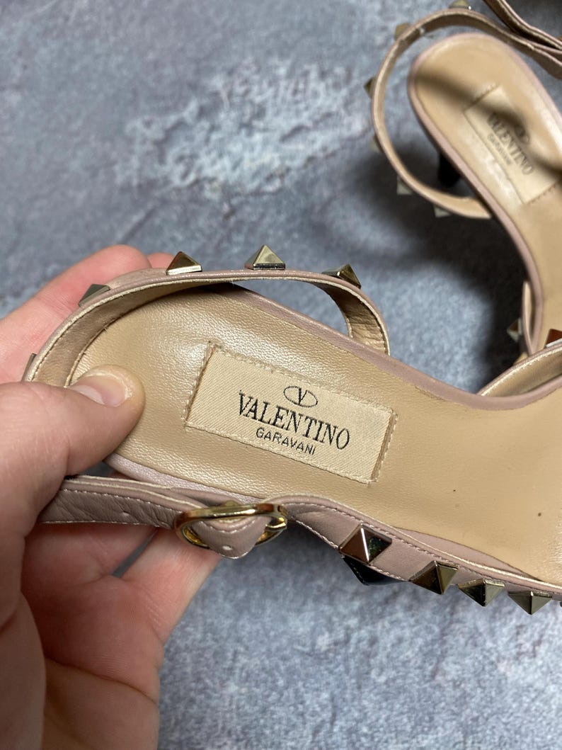 May include: Close-up of a pair of light pink Valentino Garavani sandals. The sandals feature small gold-colored pyramid studs and a thin ankle strap. The Valentino logo is visible on the insole.