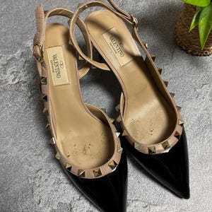 May include: A pair of black patent leather slingback heels with pointed toes. The shoes have a nude-colored trim and are adorned with gold-colored pyramid studs. The brand name "Valentino Garavani" is visible on the insole.