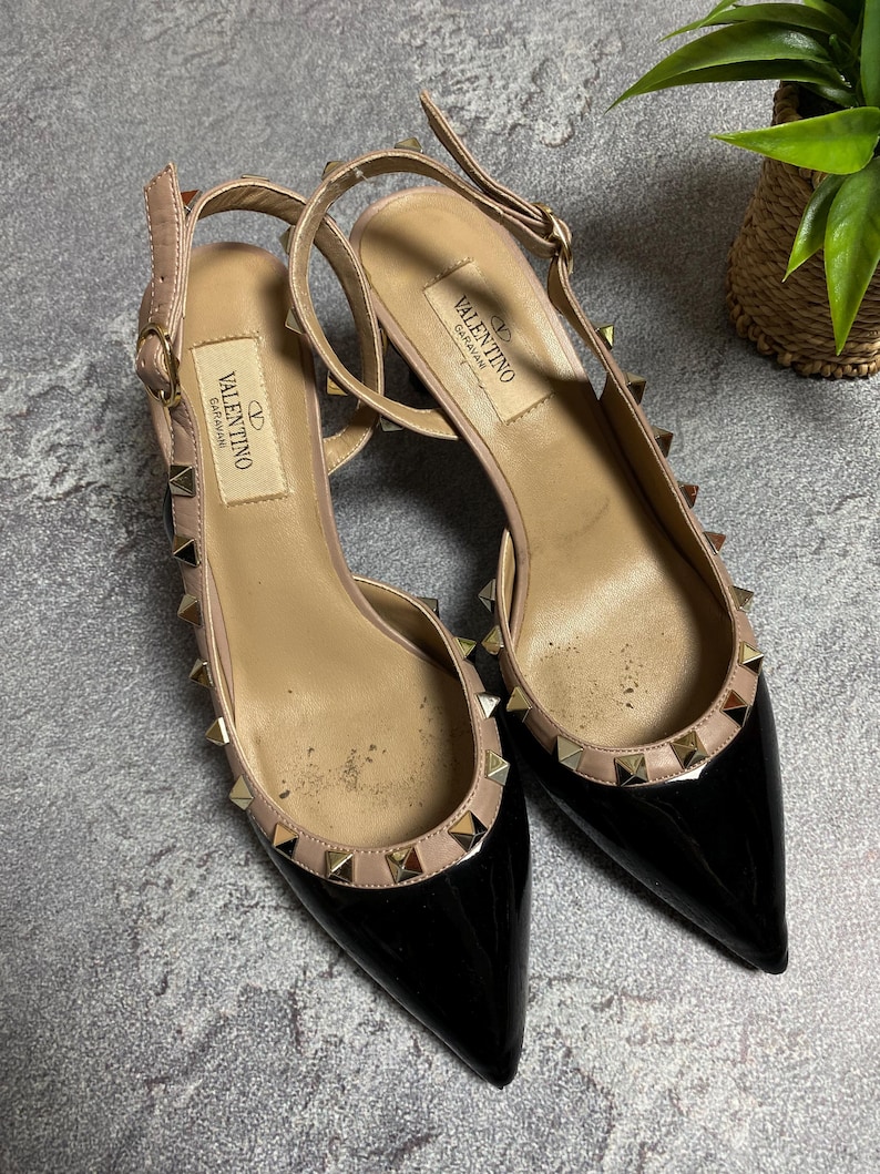 May include: A pair of black patent leather slingback heels with pointed toes. The shoes have a nude-colored trim and are adorned with gold-colored pyramid studs. The brand name "Valentino Garavani" is visible on the insole.