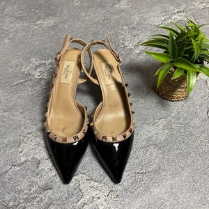 May include: A pair of black patent leather slingback heels with pointed toes. The shoes have a nude-colored insole and ankle strap, adorned with gold-tone studs along the trim. The Valentino logo is visible on the insole.