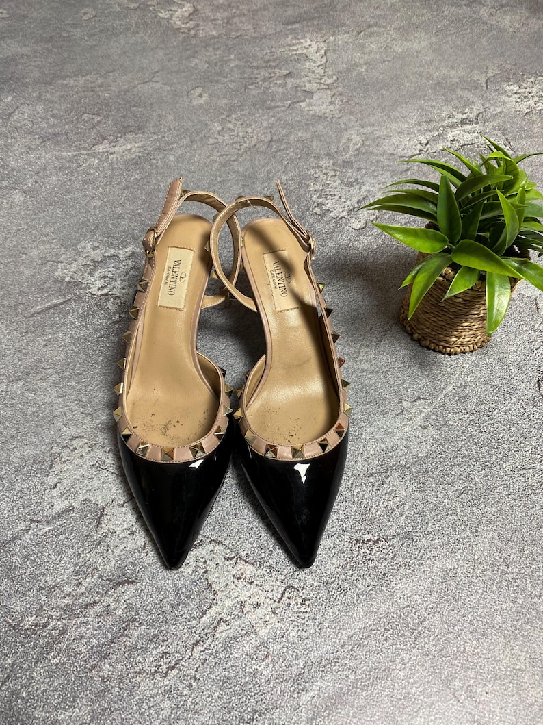 May include: A pair of black patent leather slingback heels with pointed toes. The shoes have a nude-colored insole and ankle strap, adorned with gold-tone studs along the trim. The Valentino logo is visible on the insole.