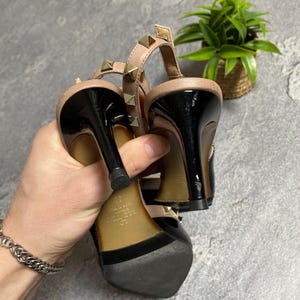 May include: Black and nude-colored high-heeled sandals with gold-colored stud accents. The sandals have a black heel and a strap around the ankle. The shoes are held up by a hand.