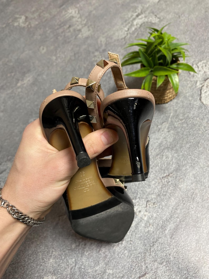 May include: Black and nude-colored high-heeled sandals with gold-colored stud accents. The sandals have a black heel and a strap around the ankle. The shoes are held up by a hand.