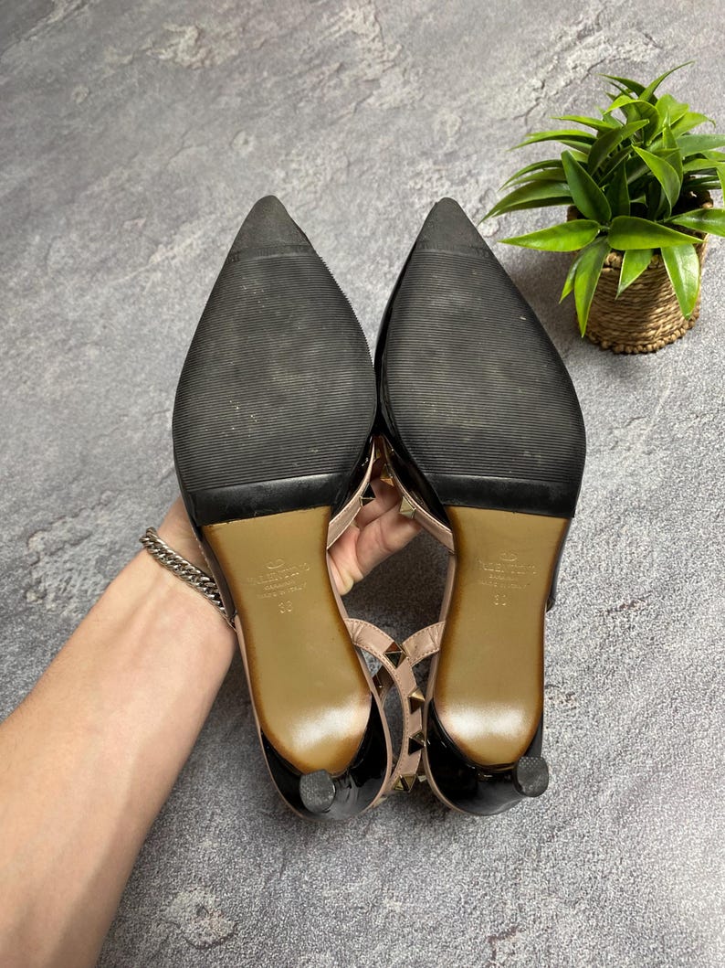 May include: Pair of black and nude-colored high-heeled shoes with pointed toes. The soles are black with a textured pattern. The heels are short and rounded. The shoes have a strap with gold-colored studs.