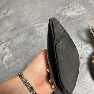 May include: Black patent leather high heel shoe with gold-tone studs. The shoe has a pointed toe and a stiletto heel. The sole is black rubber. A silver chain bracelet is visible in the image.