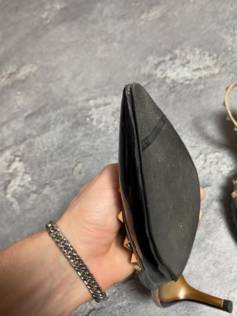 May include: Black patent leather high heel shoe with gold-tone studs. The shoe has a pointed toe and a stiletto heel. The sole is black rubber. A silver chain bracelet is visible in the image.