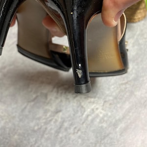 May include: Black high-heeled sandals with a glossy finish. The heels have a stiletto design. The sandals feature gold-colored stud accents and a strap design. The soles are a light brown color.