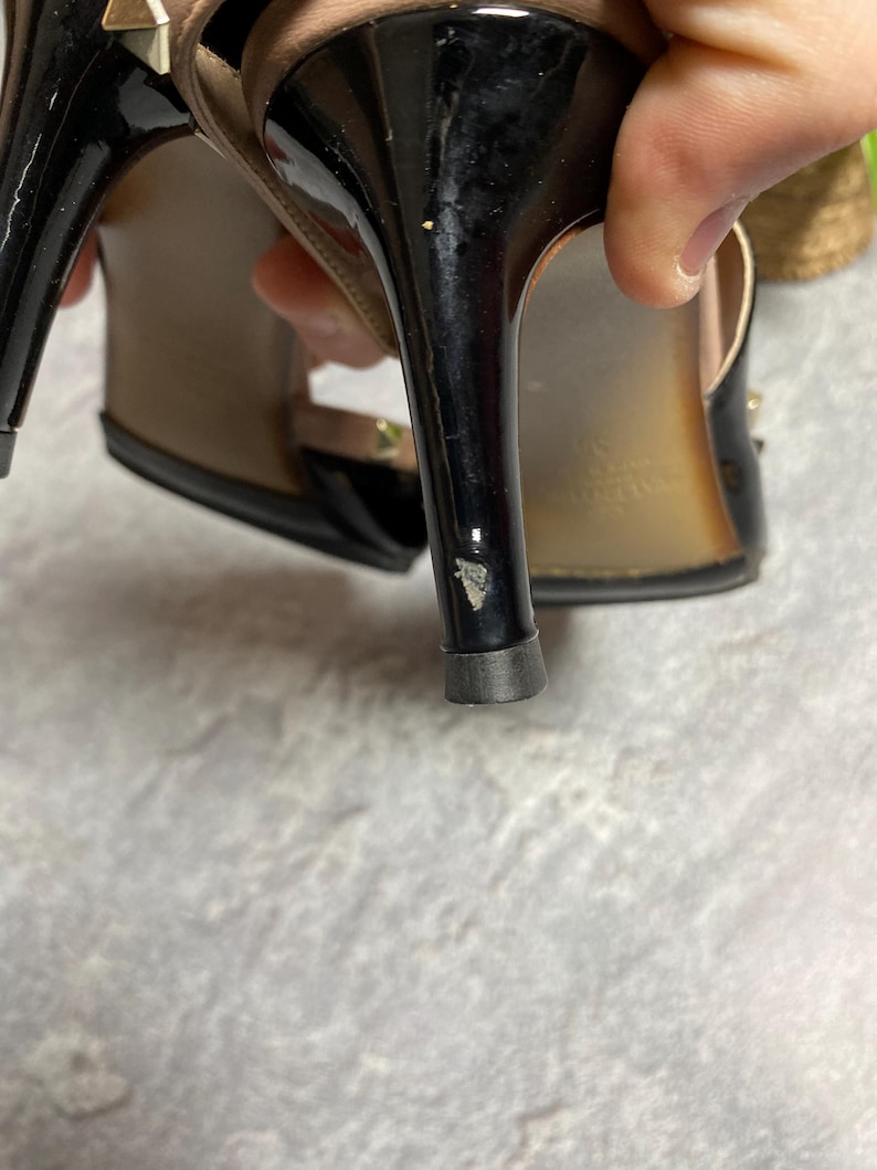 May include: Black high-heeled sandals with a glossy finish. The heels have a stiletto design. The sandals feature gold-colored stud accents and a strap design. The soles are a light brown color.