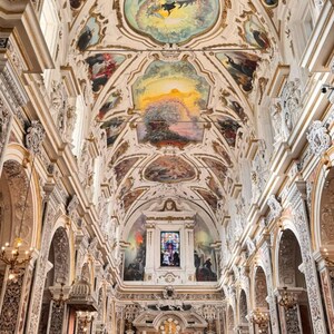 May include: Interior view of Chiesa Del Gesu Casa Profesa in Sicily. The church features ornate architectural details, including painted ceilings, arched doorways, and decorative columns. The space is illuminated by natural light and chandeliers.