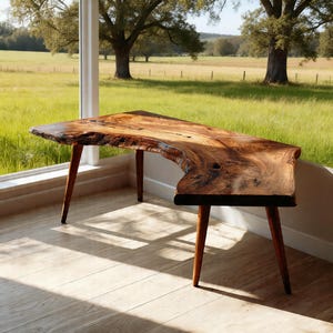 Puede incluir: Un escritorio de madera con borde natural, que muestra la forma natural de la madera. El escritorio es de color marrón oscuro, con vetas visibles y está sostenido por tres patas anguladas. El escritorio está cerca de una ventana con vista a un campo verde y árboles. El escritorio mide aproximadamente 152 cm de largo.