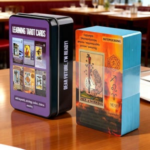 Op de afbeelding: Een tarotkaartspel met een zwarte metalen doos. Op de doos staat "LEARNING TAROT CARDS" en "DEAR FUTURE, I'M READY!". De set bevat geïllustreerde kaarten met trefwoorden. De set ligt op een houten tafel.