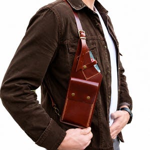 Può includere: Una borsa a tracolla in pelle marrone con un design verticale. La borsa ha una chiusura con patta con due bottoni color ottone e una lunga tracolla regolabile. La borsa è indossata a tracolla e la pelle ha una finitura ricca e lucida.