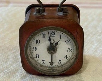 Könnte beinhalten: Ein kleiner, kupferfarbener Reise-Wecker. Die Uhr hat ein weißes Zifferblatt mit schwarzen Ziffern und Zeigern. Sie hat einen Griff oben und steht auf einer weißen Oberfläche.