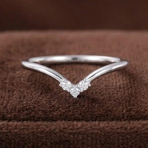 May include: A silver-toned V-shaped chevron ring featuring three small, clear, round-cut stones set at the center point. The band has a smooth, polished finish and a delicate, curved silhouette that tapers toward the back of the ring.