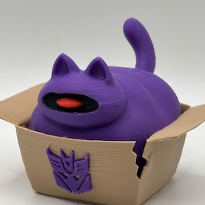 May include: A purple cat-shaped figurine with a black eye and red center, sitting in a tan cardboard box. The box features a purple emblem on the side. The cat has a curved tail and pointed ears.