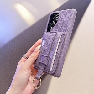 May include: A lavender phone case with a textured finish, featuring a cardholder and a gold-colored ring. The case has a camera cutout and a strap. The cardholder is designed to hold cards.