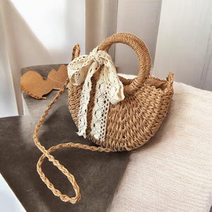 Op de afbeelding: Een geweven strooien handtas met een afgeronde vorm en een ronde handgreep. De tas heeft een crèmekleurige kanten sjaal om de handgreep geknoopt. Een bijpassende strohoed is zichtbaar op de achtergrond. De tas heeft een lange, geweven riem.