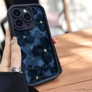 May include: Black phone case with a floral design. The case has a dark blue background with black flower patterns and gold accents. The phone case has a black border and a camera lens protector.