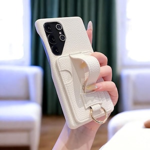 May include: A white textured phone case with a strap and a gold ring. The case has a camera cutout and a small pocket. The strap is designed for holding the phone securely. The case is made of a durable material.