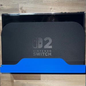 May include: Black Nintendo Switch console with a blue stand. The console features the Nintendo Switch logo and the number 2. The stand is a vibrant blue and is positioned at the bottom of the console.