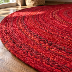 Puede incluir: Una alfombra redonda trenzada que presenta una mezcla vibrante de fibras rojas, naranjas y marrón oscuro. El diseño circular está construido con hebras textiles gruesas y estrechamente tejidas que crean un patrón concéntrico y texturizado.
