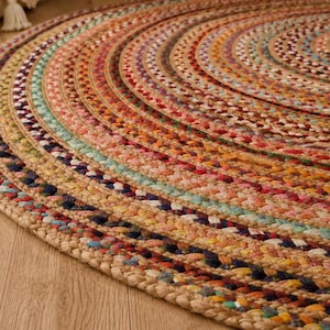May include: A round braided rug featuring concentric circles of multi-colored fabric strips woven with natural tan jute fibers. The design incorporates a vibrant palette of red, blue, yellow, green, and orange tones arranged in a repeating pattern.