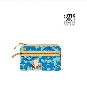 May include: A rectangular zipper pouch featuring a blue floral pattern with a small bird illustration. The pouch has two tan zippered compartments with orange pull tabs and the text ZIPPER POUCH SEWING PATTERN printed in black above the item.