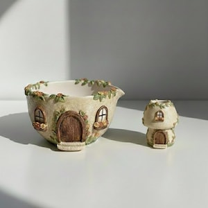 Puede incluir: Una maceta de cerámica beige caprichosa y un pequeño portavelas con forma de hongo. La maceta tiene forma de casa con puerta y ventanas de madera, adornada con detalles florales. El portavelas refleja el diseño de la casa.