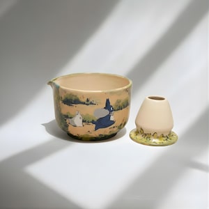 May include: A beige ceramic bowl with a spout and a small, matching cream-colored container. The bowl features a painted scene with cartoon characters in a landscape. The container sits on a yellow and green base.