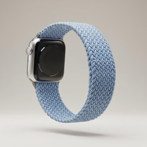 May include: A silver smartwatch with a black screen and a light blue braided band. The band is made of a woven material and is designed to fit around the wrist. The watch is shown against a neutral background.