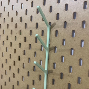 May include: A light green pegboard hook with three arms, designed to hang items. The hook is mounted on a brown pegboard with evenly spaced oval holes. The hook is made of plastic.
