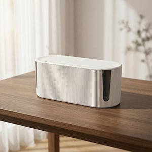 May include: A white cable tidy box with a ribbed design and a removable lid. The box has rounded edges and dark grey openings at each end. It rests on a dark brown wooden table.