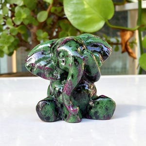 May include: A carved stone elephant figurine featuring a polished surface with a mottled pattern of deep forest green and vibrant magenta red. The sculpture depicts a seated elephant with large ears, a curved trunk, and visible eye details.