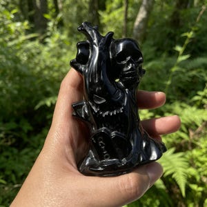May include: A polished black stone carving featuring a detailed skull positioned above a gnarled tree trunk. The base of the sculpture includes a small tombstone with the letters RIP clearly engraved on the front surface of the dark material.